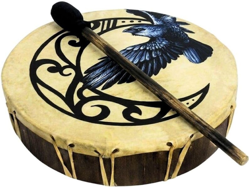 May include: A round frame drum with a light tan drumhead featuring a black graphic of a crescent moon and a flying raven. The drum has a dark wooden frame laced with tan leather strips and includes a wooden mallet with a black felt head.