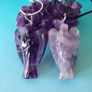 May include: Two amethyst angel pendants on chains. One pendant is on a silver chain and the other is on a black cord. The pendants are carved from amethyst crystals and have a purple and white color.