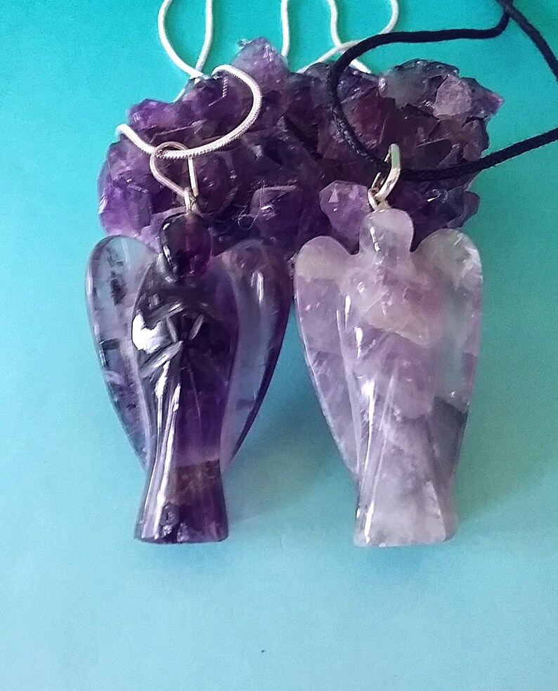 May include: Two amethyst angel pendants on chains. One pendant is on a silver chain and the other is on a black cord. The pendants are carved from amethyst crystals and have a purple and white color.