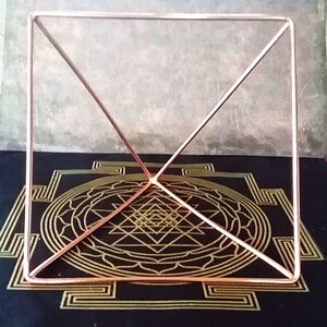 K&ouml;nnte beinhalten: Ein Kupfer-Pyramidenrahmen auf einer schwarzen Oberfl&auml;che mit einem goldenen geometrischen Muster. Die Pyramide besteht aus d&uuml;nnem Kupferdraht und hat eine quadratische Basis.