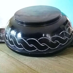 May include: A black, round, carved stone bowl with a decorative wave pattern. The bowl is made of soapstone and has a smooth, polished finish.