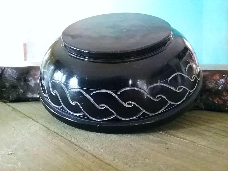 May include: A black, round, carved stone bowl with a decorative wave pattern. The bowl is made of soapstone and has a smooth, polished finish.