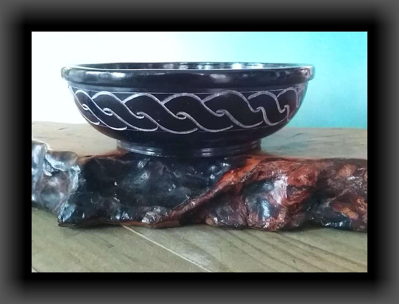 May include: A black slate bowl with a carved white design. The bowl is sitting on a piece of wood.
