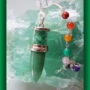 May include: A green crystal pendulum with a silver-colored metal cap and accents. The pendulum is attached to a chain with a series of colorful beads, including red, orange, yellow, green, pink, black, and purple. The background is a light green, textured surface.