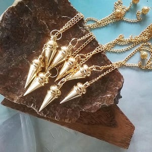 Peut inclure: Plusieurs pendules dorés avec un design conique et des chaînes attachées. Les pendules sont présentés sur une surface texturée brun foncé. Les chaînes ont de petits accents de boules décoratives.