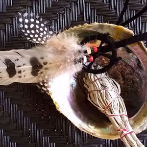 Puede incluir: Una concha de abulón blanca con un manojo de varitas de salvia blanca atadas con cuerda roja y una pluma moteada de blanco y negro unida a un cordón negro con cuentas rojas.