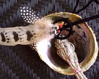 May include: A white abalone shell with a bundle of white sage smudge sticks tied with red string and a black and white spotted feather attached to a black cord with red beads.