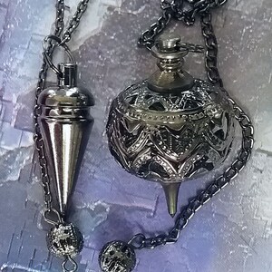 May include: Two silver-colored pendulums hanging from chains. One is a cone shape, the other is a decorative sphere with an openwork design. Both have small, textured spheres at the end of their chains. The background is a soft, mottled purple and white.