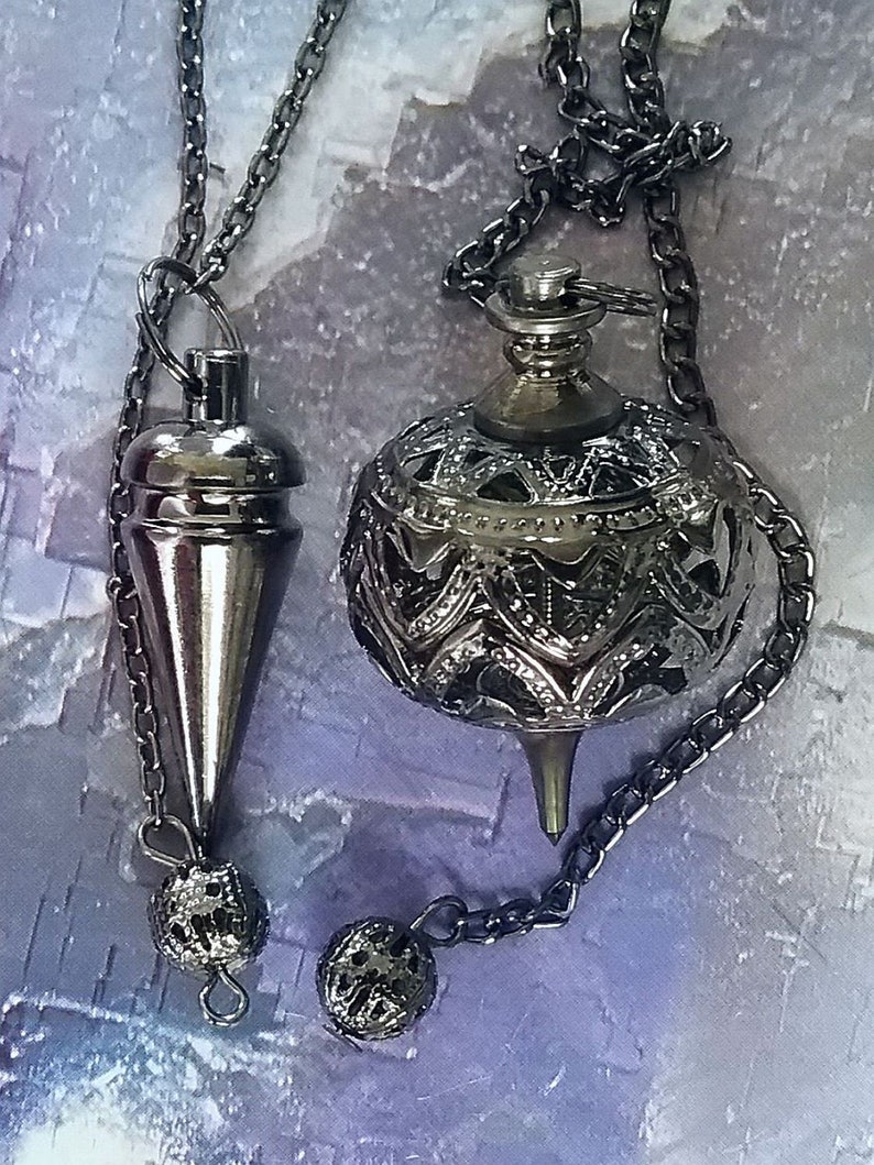 May include: Two silver-colored pendulums hanging from chains. One is a cone shape, the other is a decorative sphere with an openwork design. Both have small, textured spheres at the end of their chains. The background is a soft, mottled purple and white.