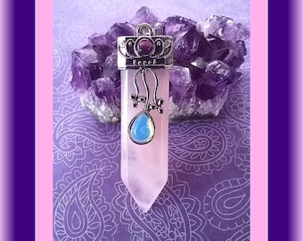 May include: A pointed, pale pink crystal pendant featuring a silver-toned metal cap with a small, round, dark purple stone and a dangling, teardrop-shaped, iridescent blue stone accent, suspended in front of a cluster of deep purple amethyst.