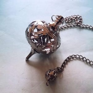 May include: A silver-colored pendulum with a chain. The pendulum has a spherical, openwork design and a pointed tip. A smaller, matching sphere hangs at the end of the chain. The background is a light blue.