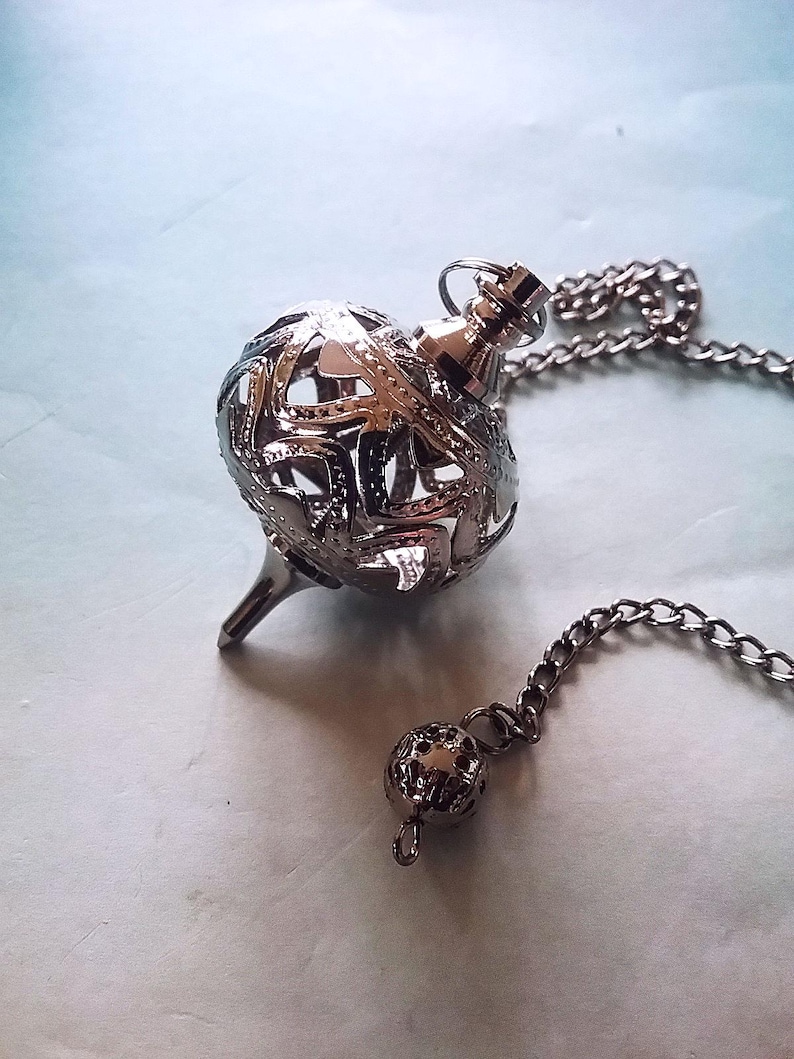 May include: A silver-colored pendulum with a chain. The pendulum has a spherical, openwork design and a pointed tip. A smaller, matching sphere hangs at the end of the chain. The background is a light blue.
