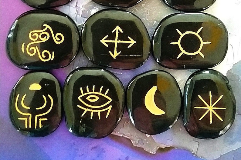 May include: Eight black smooth stones with gold symbols. The symbols include a crescent moon, a sun, a star, an eye, a swirl, a cross with arrows, a tree, and a starburst.