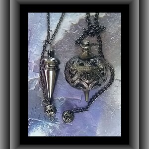 May include: Two silver-toned pendulums hanging from chains. One is a cone shape, the other is a decorative sphere with intricate cut-out designs. Both have small, decorative ball-shaped weights at the bottom. The background is a textured, light blue and purple.
