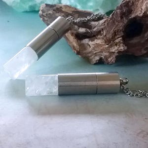 Può includere: Due collane con pendente color argento con inserti in cristallo trasparente e rettangolare. I pendenti sono su delicate catene d'argento. Lo sfondo comprende un pezzo di legno alla deriva e una superficie azzurra.