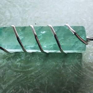May include: A light green crystal pendant wrapped in silver wire. The crystal is faceted and has a smooth, polished finish.