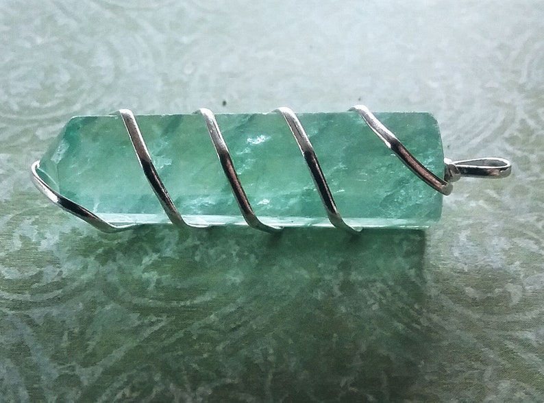 May include: A light green crystal pendant wrapped in silver wire. The crystal is faceted and has a smooth, polished finish.