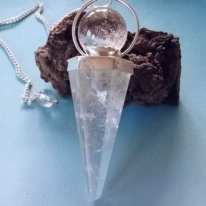 Puede incluir: Un péndulo de cristal de cuarzo transparente con una cadena de plata y una esfera de vidrio transparente en la parte superior. El péndulo descansa sobre una roca marrón oscura.