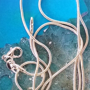 Op de afbeelding: Een zilveren slangenketting met een karabijn sluiting. De ketting ligt op een blauwe en groene achtergrond.