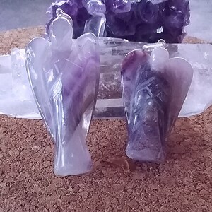 May include: Two amethyst angel pendants, one with a lighter purple hue and the other with a darker purple hue, are displayed on a white crystal surface.