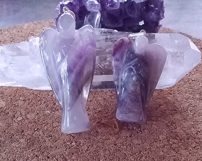May include: Two amethyst angel pendants, one with a lighter purple hue and the other with a darker purple hue, are displayed on a white crystal surface.