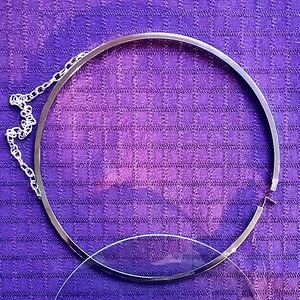 Op de afbeelding: Een zilverkleurige, ronde brillenhouder met een ketting en een heldere glazen lens. De houder ligt op een paarse geruite stof.