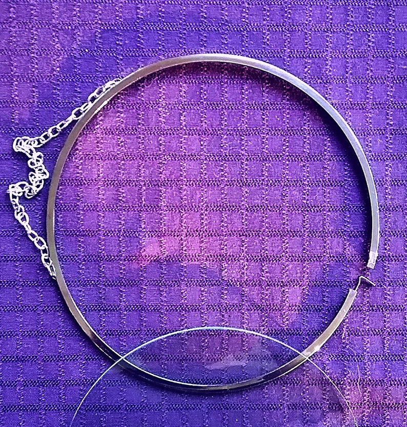 Op de afbeelding: Een zilverkleurige, ronde brillenhouder met een ketting en een heldere glazen lens. De houder ligt op een paarse geruite stof.