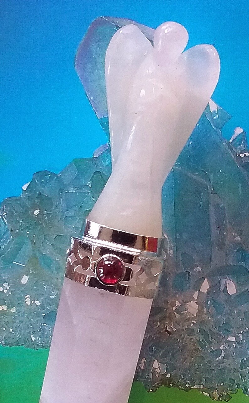 May include: White angel shaped crystal wand with a silver band and a red gemstone. The wand is sitting on a bed of blue crystals.