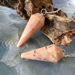 Op de afbeelding: Twee perzikkleurige, gefacetteerde, kegelvormige stenen hangers, elk bevestigd aan een zilverkleurige metalen ketting. De stenen vertonen natuurlijke variaties in kleur en textuur, waarbij de ene verticaal en de andere horizontaal is geplaatst.