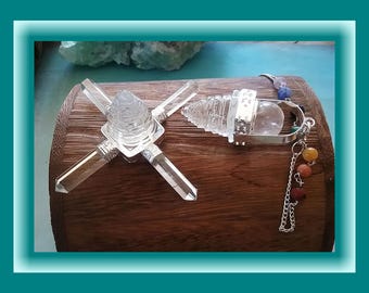 Puede incluir: Dos péndulos de cristal transparente con detalles plateados descansan sobre una superficie de madera. Uno tiene un diseño en forma de cruz con puntas de cristal. El otro presenta una bola de cristal transparente y una cadena con cuentas de colores. El fondo incluye un borde turquesa.