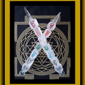 Pode incluir: Duas varinhas de quartzo transparente com letras vermelhas e azuis, cruzadas uma sobre a outra em um fundo preto com um design de mandala dourado. As varinhas são adornadas com contas de quartzo transparente.