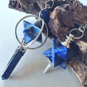 May include: Two pendants featuring deep blue lapis lazuli gemstones. One pendant has a star-shaped stone inside a silver-toned metal ring with a pointed crystal base. The second pendant features a star-shaped lapis lazuli stone with a silver-toned cap.
