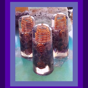 Op de afbeelding: Drie doorzichtige glazen flessen gevuld met amethistkristallen en koperen draadspiralen. De flessen zijn in een rij op een blauw-groen oppervlak gerangschikt.