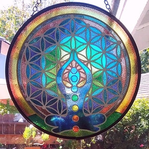 Op de afbeelding: Een hangend glas-in-loodraam met een kleurrijk mandala-ontwerp met een levensbloemmotief en een blauwe yogafiguur met chakra-symbolen.