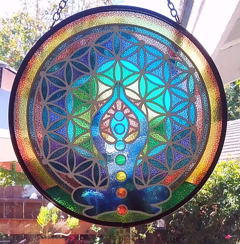 Op de afbeelding: Een hangend glas-in-loodraam met een kleurrijk mandala-ontwerp met een levensbloemmotief en een blauwe yogafiguur met chakra-symbolen.