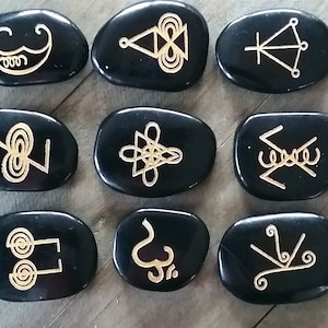 May include: Nine black oval-shaped stones with gold symbols etched on the surface. The stones are arranged in three rows of three.