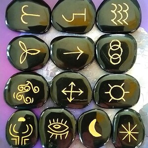 May include: Twelve black, smooth, oval-shaped stones with gold symbols etched on them. The symbols include an arrow, a crescent moon, a sun, a circle with three smaller circles inside, a cross with arrows pointing out, a spiral, a triquetra, a starburst, an eye, a wave, and the astrological signs for Aries, Taurus, and Gemini.