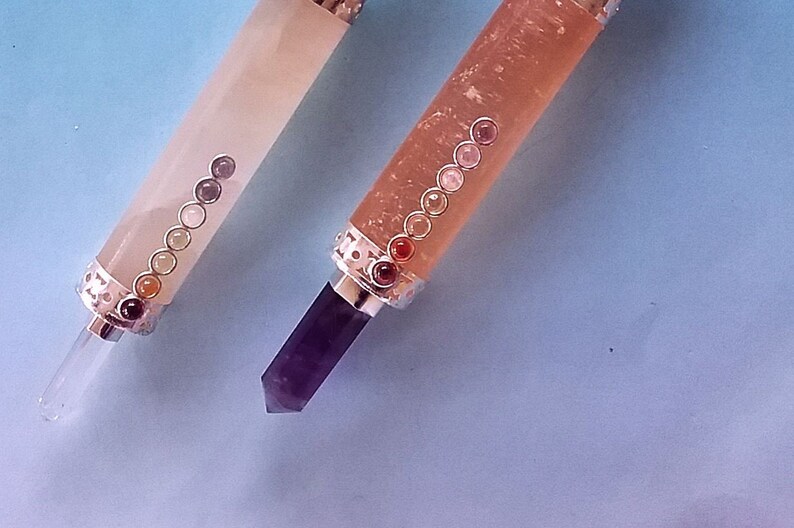 May include: Two crystal wands with silver detailing and coloured stones. One wand is pale pink with a clear crystal tip, the other is a darker pink with a purple crystal tip. The wands have a series of small, coloured stones.