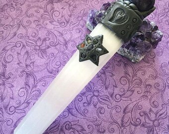 May include: A white cylindrical selenite wand featuring a dark gray clay cap with a heart motif and a dark gray star-shaped embellishment inlaid with small multicolored stones. A dark sphere sits atop the wand, which rests against a cluster of purple amethyst crystals.