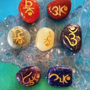 May include: Seven smooth, polished stones, each a different colour, with gold Sanskrit symbols inscribed on them. The stones are arranged in a circle on a blue and green background.