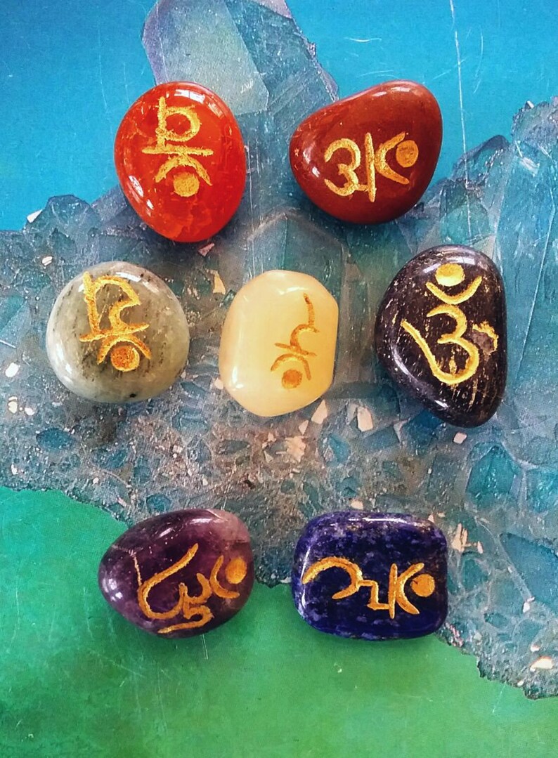 May include: Seven smooth, polished stones, each a different colour, with gold Sanskrit symbols inscribed on them. The stones are arranged in a circle on a blue and green background.