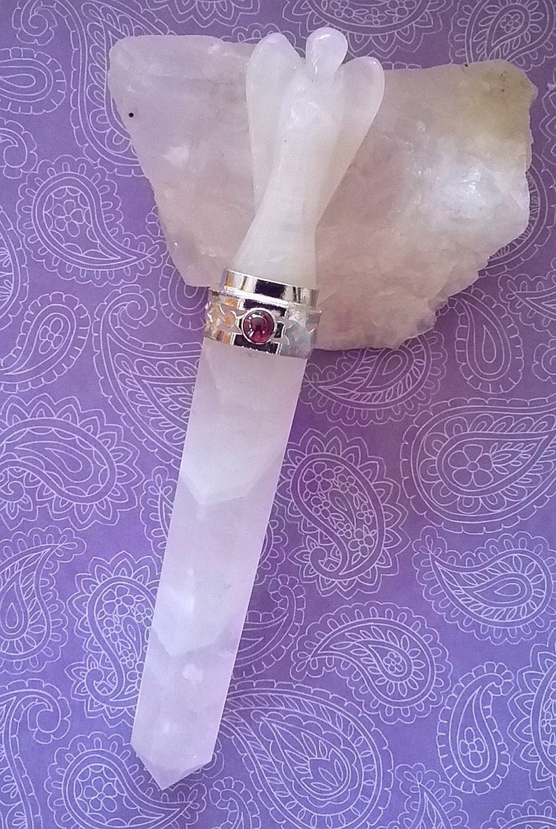 May include: A white angel wing shaped crystal wand with a silver band and a pink gemstone. The wand is resting on a piece of white crystal.
