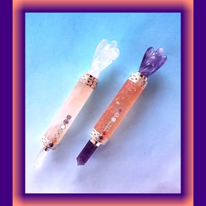 May include: Two crystal wands with angel-shaped tops. One is pale pink with a clear crystal tip and a row of coloured stones. The other is orange with an amethyst tip and a similar stone row.