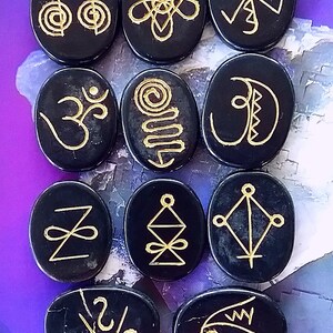 May include: Twelve black oval-shaped stones with gold symbols etched on them. The symbols include a spiral, a triangle, a crescent moon, and an infinity symbol.