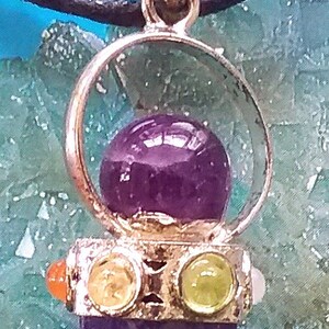 Puede incluir: Un colgante de plata con una piedra de amatista p&uacute;rpura rodeada por un c&iacute;rculo de plata. El colgante est&aacute; adornado con cuatro piedras m&aacute;s peque&ntilde;as: dos naranjas, una amarilla y una verde.
