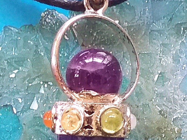 Puede incluir: Un colgante de plata con una piedra de amatista p&uacute;rpura rodeada por un c&iacute;rculo de plata. El colgante est&aacute; adornado con cuatro piedras m&aacute;s peque&ntilde;as: dos naranjas, una amarilla y una verde.