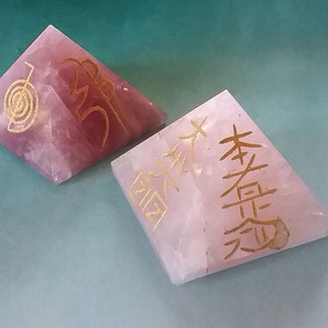 May include: Two pink crystal pyramids with gold lettering. The pyramid on the left has a spiral symbol and the word "Om" inscribed on it. The pyramid on the right has Chinese characters inscribed on it.