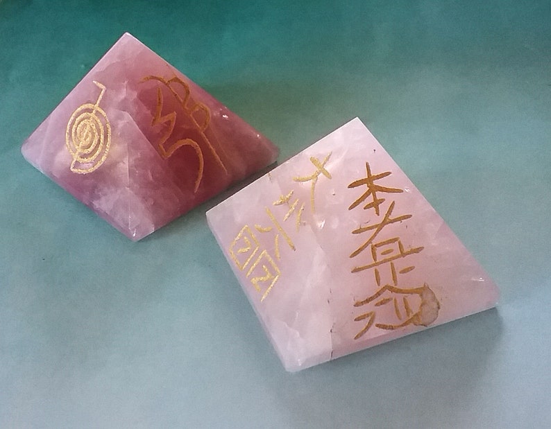 May include: Two pink crystal pyramids with gold lettering. The pyramid on the left has a spiral symbol and the word "Om" inscribed on it. The pyramid on the right has Chinese characters inscribed on it.