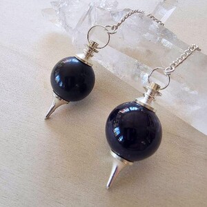 Op de afbeelding: Twee bolvormige zwarte stenen pendels met zilverkleurige metalen doppen en puntige uiteinden. Elke pendel is bevestigd aan een dunne zilverkleurige metalen ketting. De pendels zijn geplaatst voor een helder, langwerpig, grillig kwartskristal.