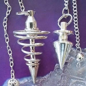 May include: Two silver-colored pendulums with chains hang against a purple background. One pendulum has a spiral design, and the other has a conical shape. The chains are attached to the top of each pendulum and have decorative elements at the end.